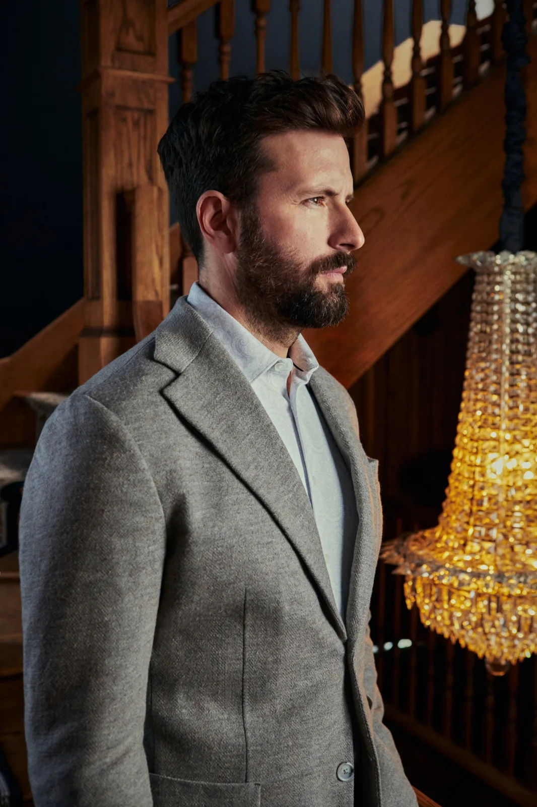 De profil en veston gris, près d’un lustre de cristal.